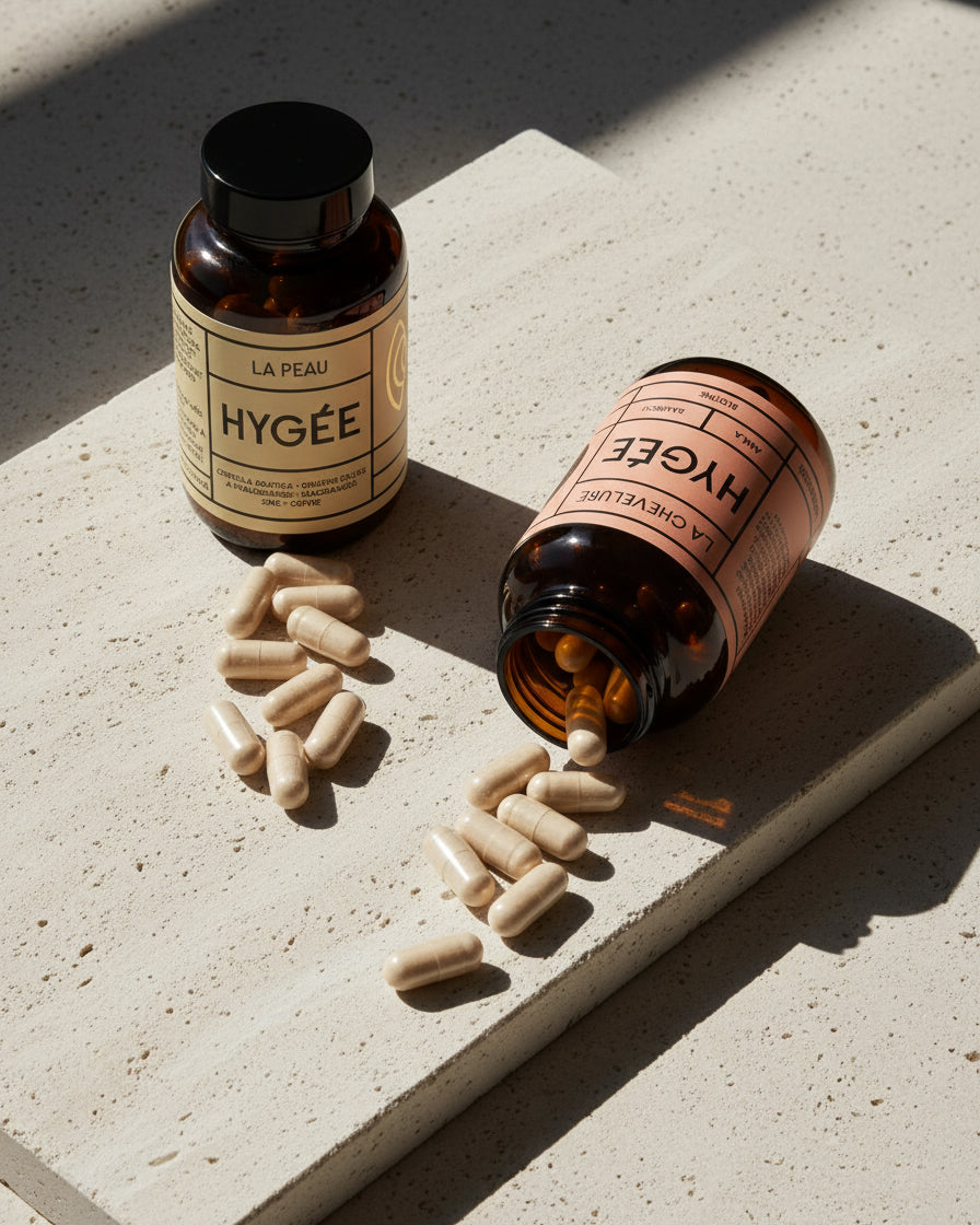 La Peau stands upright and La Chevelure rests on its side with beige capsules spilling across a sunlit travertine slab, highlighting the amber jars and clean minimalist styling.