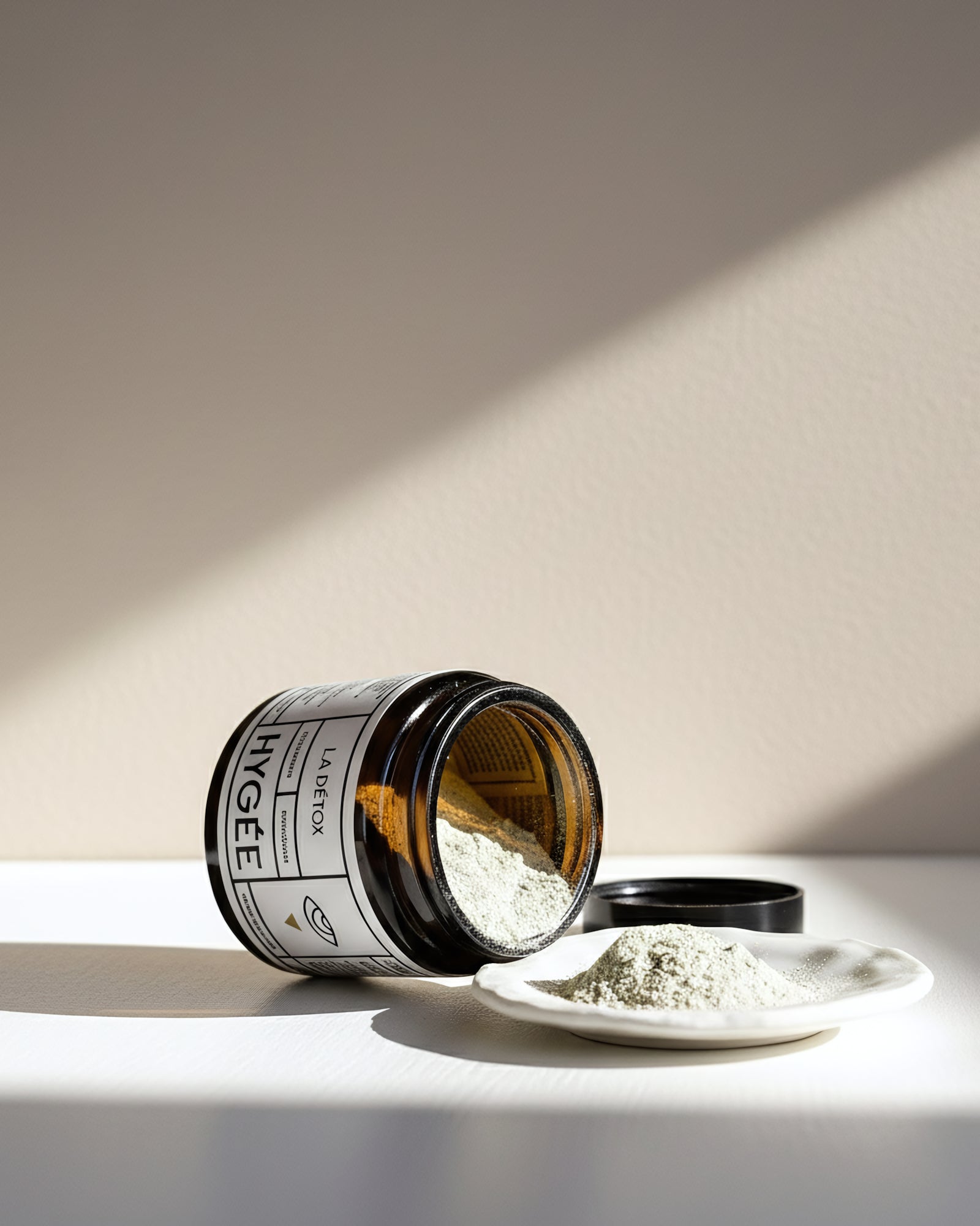 An amber HYGÉE La Détox jar lies on a sunlit white surface with its black lid off as pale botanical detox powder spills onto a small white ceramic dish, framed by soft diagonal shadows on a warm beige wall.