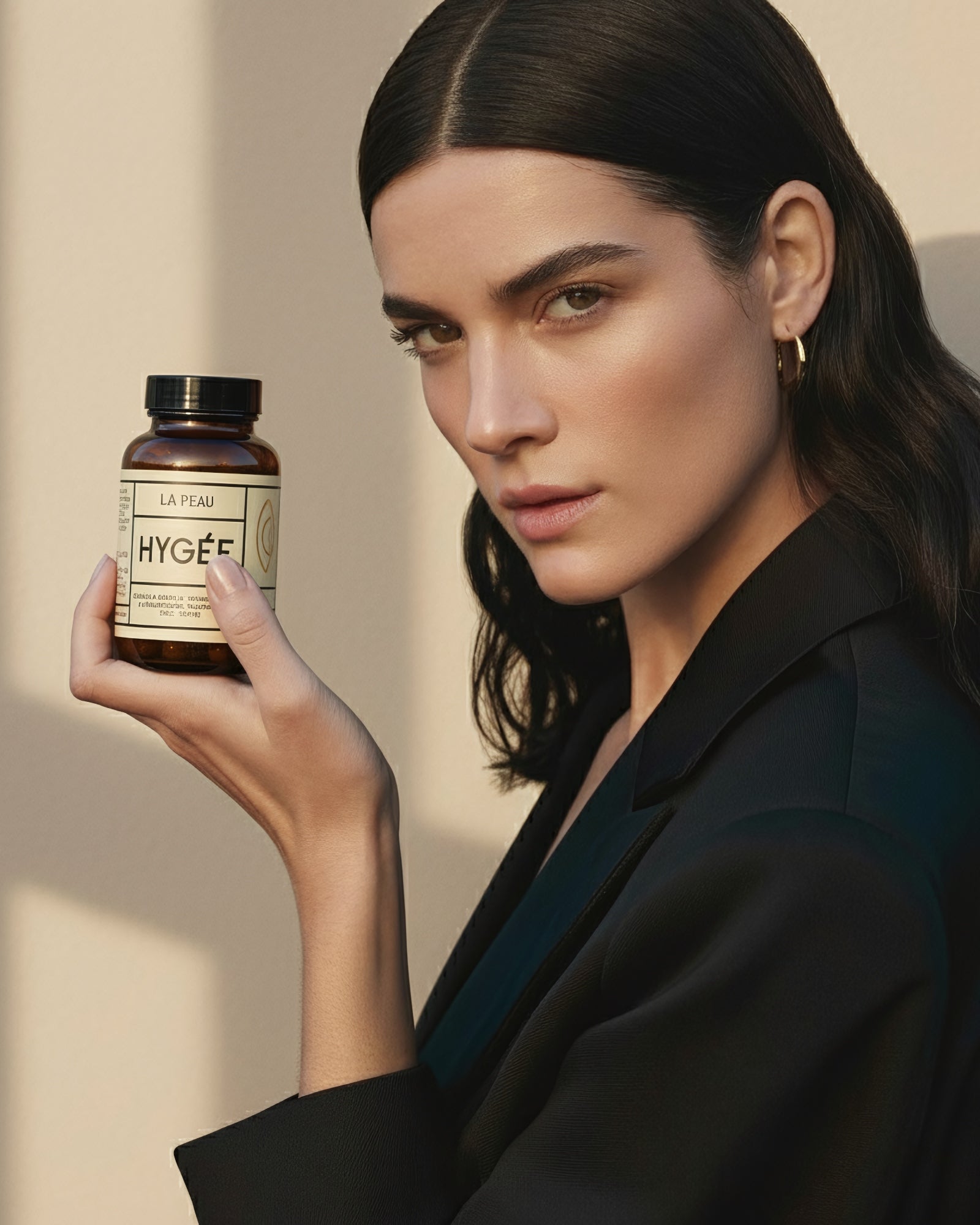 A woman in a tailored black blazer holds Allnord’s HYGÉE La Peau bottle near her face against a sunlit beige wall. A confident lifestyle portrait with clean lines and soft, directional shadow.