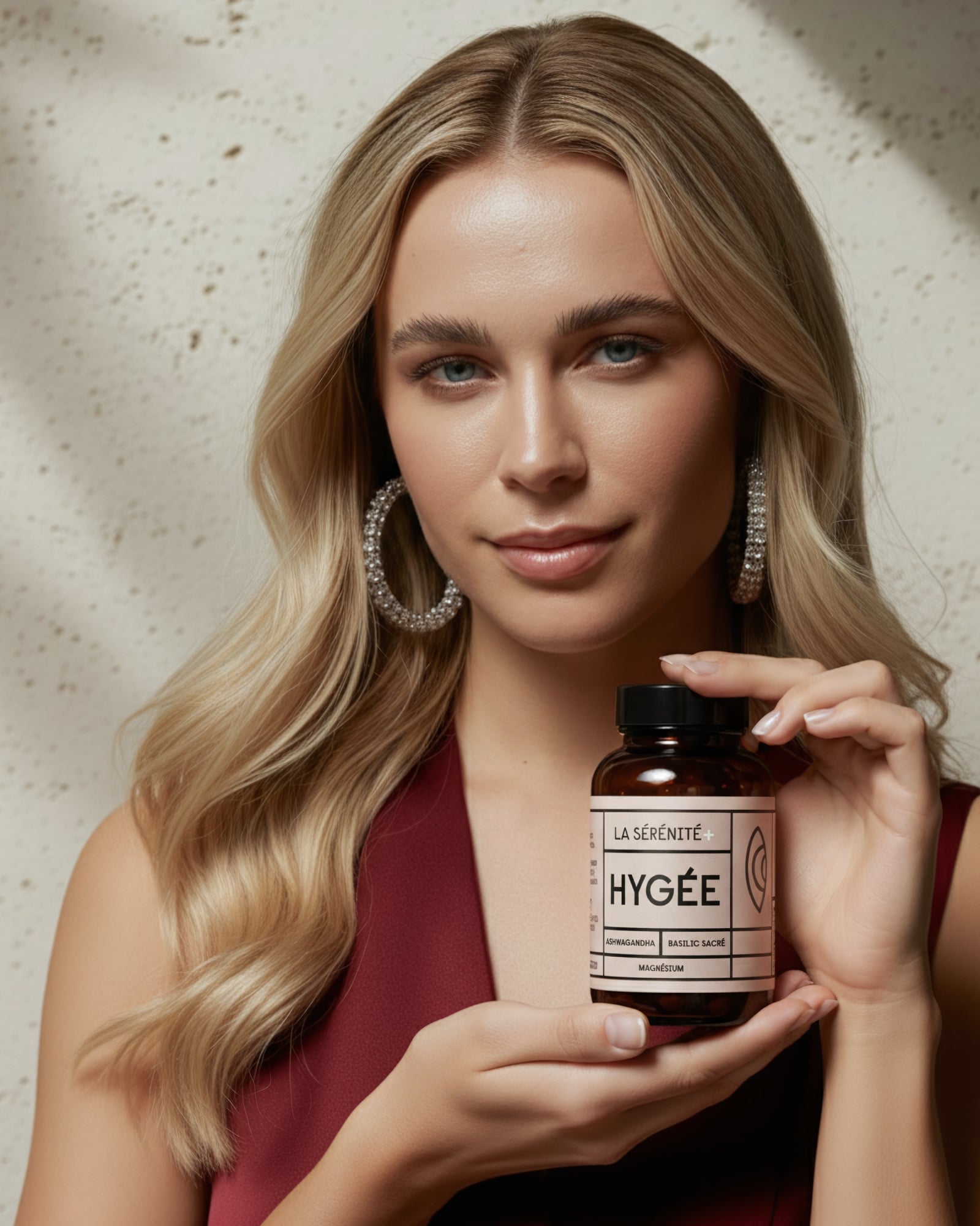A person with wavy blonde hair holds an amber HYGÉE La Sérénité+ bottle at chest height against a speckled light wall, with soft diagonal sunlight and gentle shadows.