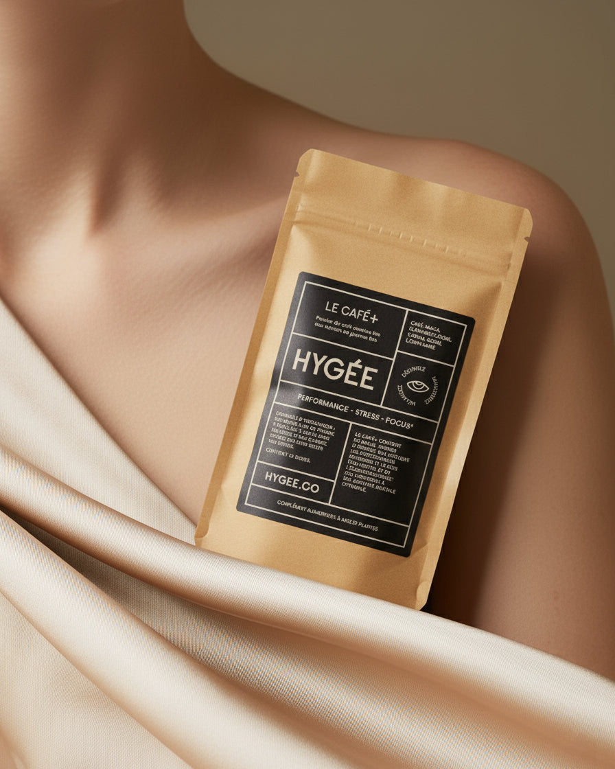 A kraft HYGÉE Le Café+ pouch with a black label rests against a bare shoulder and soft beige satin fabric, photographed in gentle studio light for a refined beauty look.