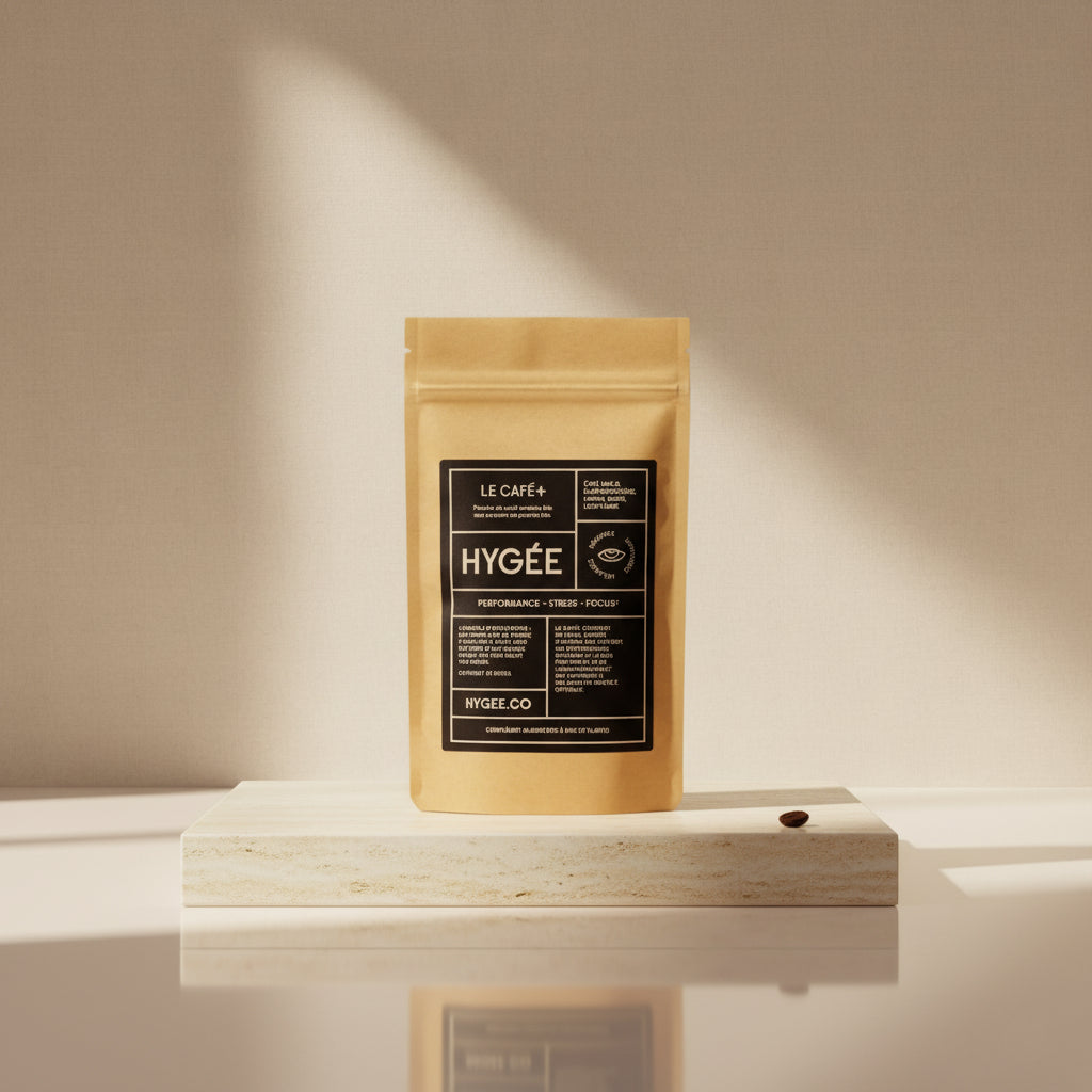 A front‑facing kraft pouch with a black HYGÉE label stands centered on a pale stone plinth, lit by a narrow beam of sunlight that creates soft wall shadows and a faint base reflection. A minimalist studio portrait in creamy neutrals.