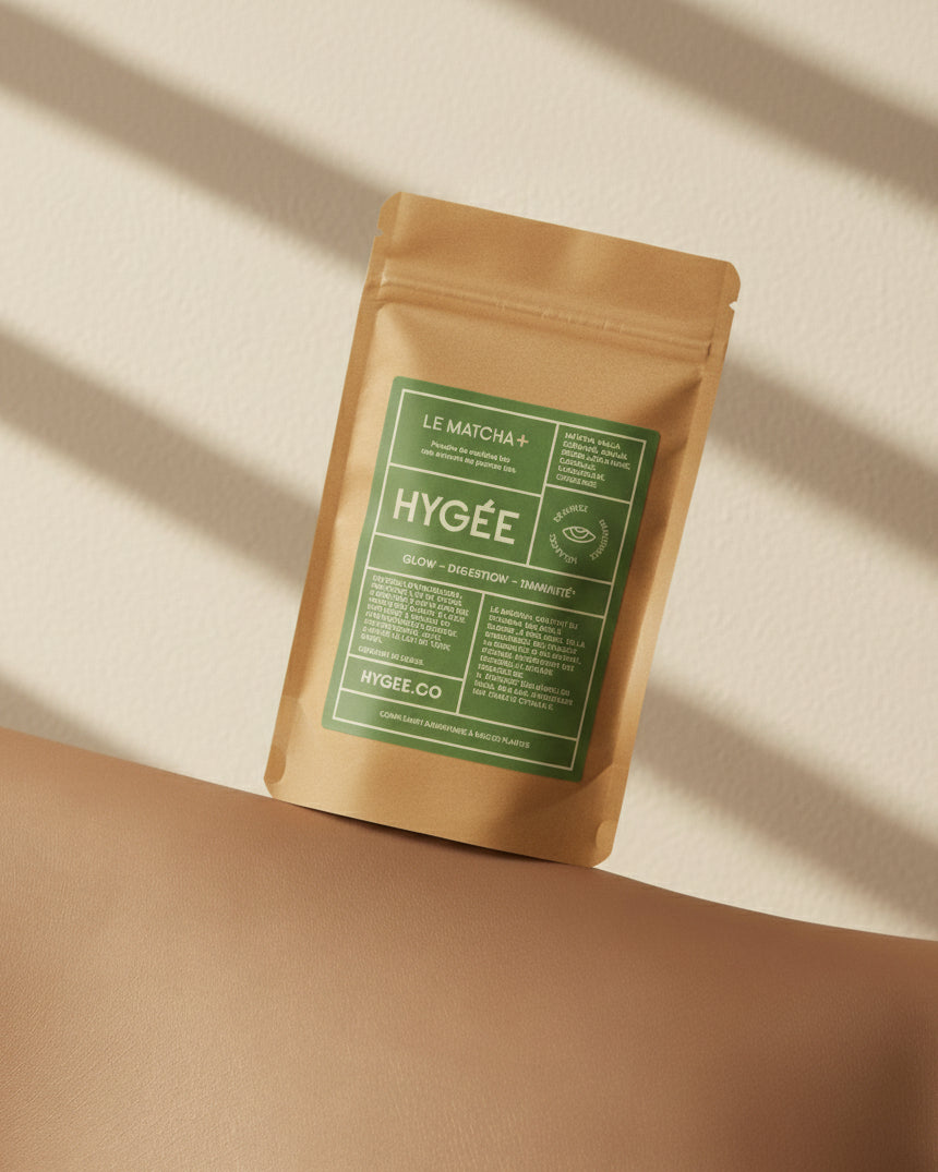 A kraft HYGÉE Le Matcha+ pouch with a green label balances on a smooth tan curve against a textured off‑white wall, crossed by soft linear shadows.