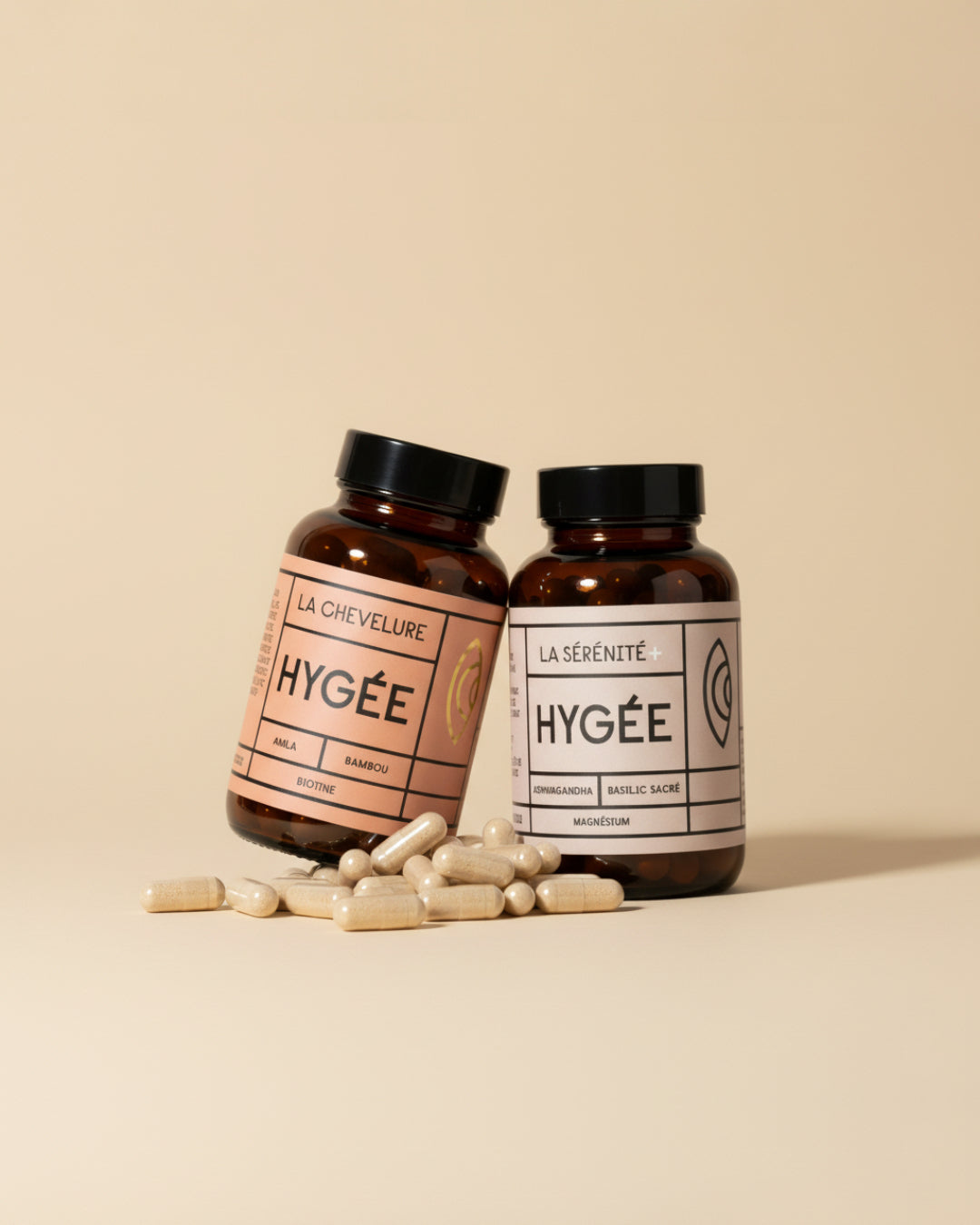 Two amber HYGÉE bottles, La Chevelure and La Sérénité+, leaning together on a soft beige backdrop with a scatter of cream capsules in the foreground, creating a warm minimal wellness scene.