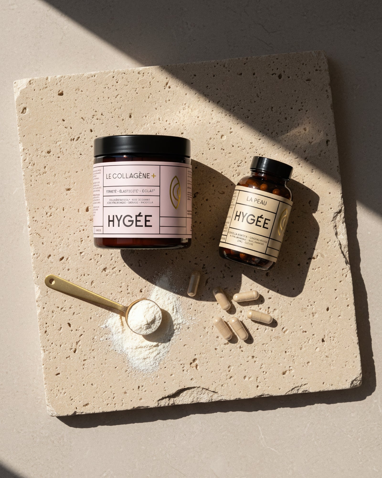 Overhead flatlay showing HYGÉE Le Collagène+ jar and La Peau skin capsules on a textured stone slab with a golden spoon of collagen powder and scattered capsules, illustrating daily dosage and results‑driven inner‑beauty routine.