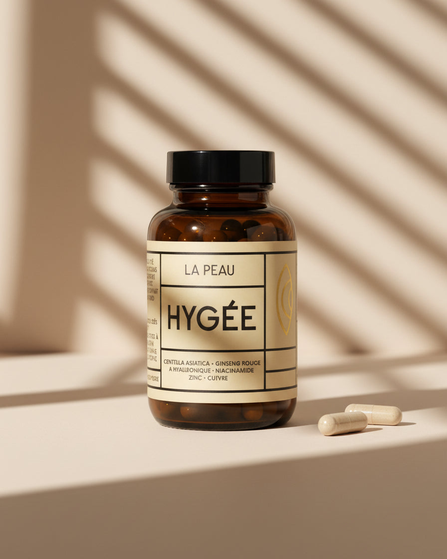 An amber glass HYGÉE La Peau bottle with a beige label stands beside two ivory capsules on a pale surface under diagonal striped shadows. A clean studio shot with sharp detail and warm neutral palette.