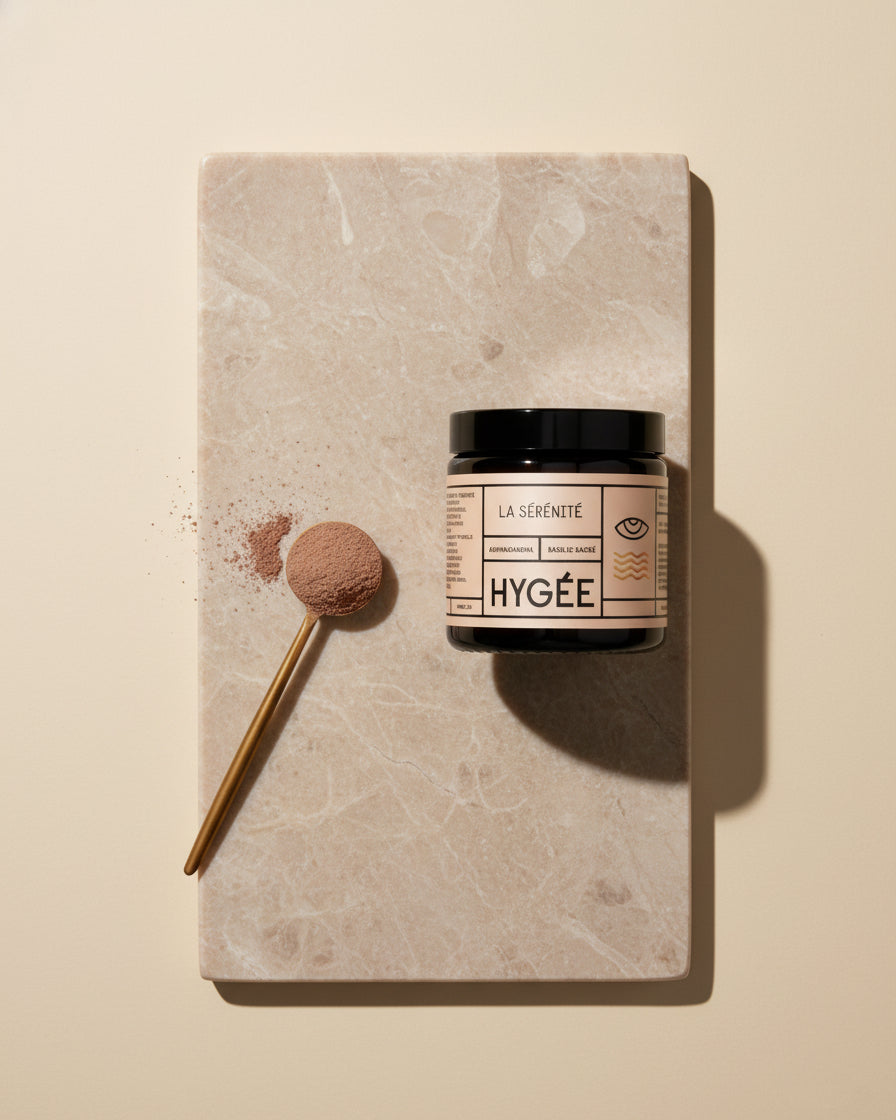 Top‑down still life of a HYGÉE La Sérénité jar on a pale marble board beside a gold measuring spoon filled with fine cocoa‑beige powder and a small spill under crisp studio light.