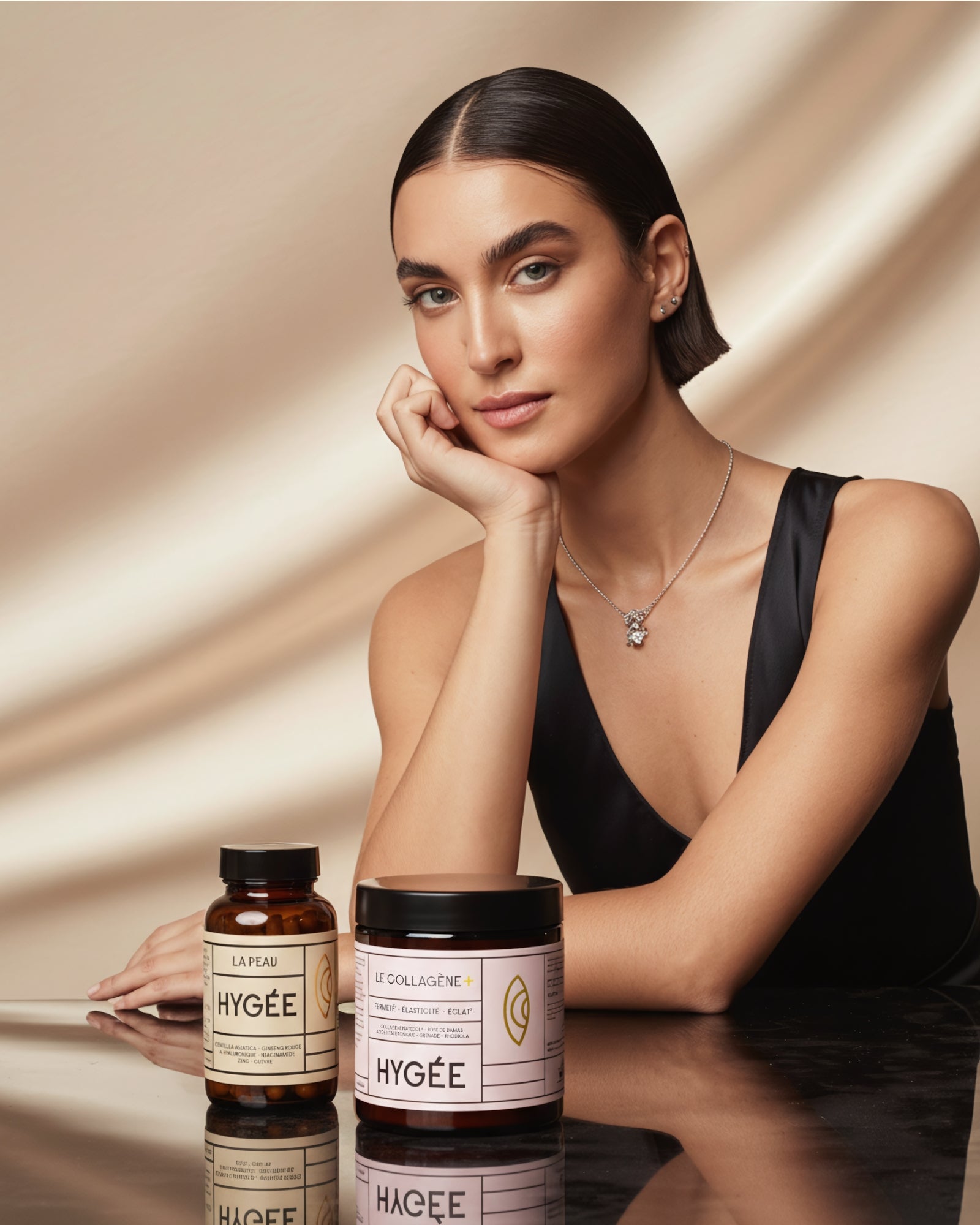 Elegant woman in a black dress sits at a glossy table beside HYGÉE Le Collagène+ collagen powder and La Peau skin capsules, evoking a premium inner‑beauty routine for firmness, radiance and complexion support.​