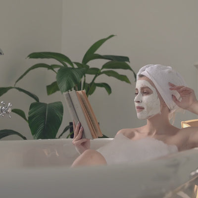 A cinematic, slow-motion shot of a woman relaxing in a bubble bath, wearing a white towel on her head and a clay face mask. She is calmly reading a book in a soft, naturally lit bathroom with lush green plants in the background, evoking a serene self-care ritual and spa-like tranquility.